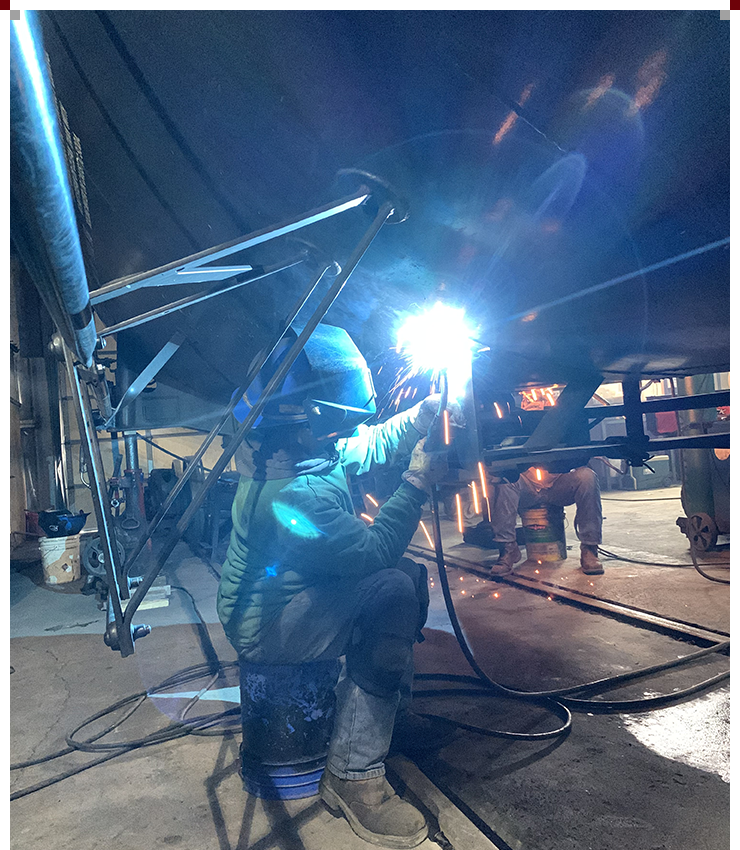 A man welding in an industrial setting with many people.